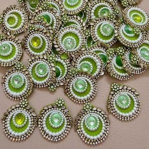May include: A collection of decorative embellishments with a circular design. Each piece features a central green jewel surrounded by white pearls, smaller green jewels, and a gold-colored border. The overall aesthetic is ornate and detailed.