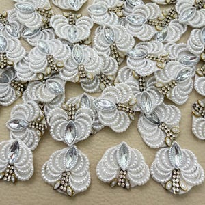 Puede incluir: Una colección de apliques florales bordados en blanco con intrincados abalorios, pequeñas perlas blancas y cristales transparentes de talla marquesa en el centro. Cada pieza tiene un diseño de pétalos simétrico con detalles en tono dorado metálico.