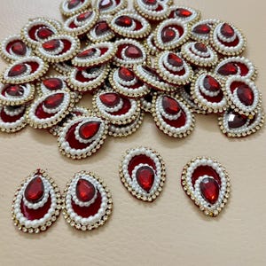 May include: Numerous red teardrop-shaped embellishments, each featuring a central red gem, encircled by white pearls and gold-toned trim. Ideal for craft projects, these decorative pieces are eye-catching.