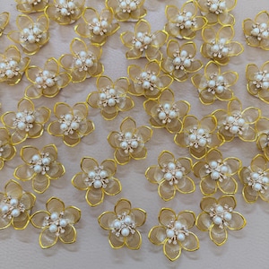 Könnte beinhalten: Eine Ansammlung zarter, goldfarbener Blumenverzierungen. Jede Blume hat fünf Blütenblätter, eine zentrale Anordnung aus Kunstperlen und funkelnden Akzenten. Das aufwendige Design deutet auf Eleganz und Detailreichtum hin, geeignet für Bastelprojekte oder Verzierungen.
