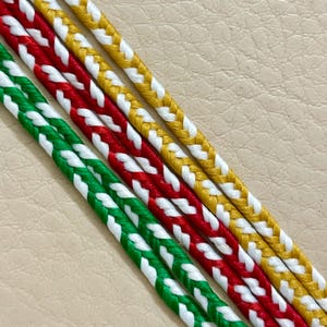 Può includere: Primo piano di diversi cordoncini intrecciati in verde, rosso e giallo, ciascuno con accenti bianchi. I cordoncini sono disposti diagonalmente, mostrando l'intricato motivo dell'intreccio e le vivaci combinazioni di colori. Lo sfondo è beige neutro.