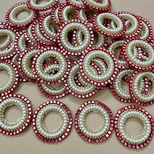 Peut inclure: Une collection d'ornements circulaires décoratifs. Chaque anneau présente une bordure extérieure rouge, un anneau central de petites perles blanches et un anneau intérieur doré. Les ornements sont disposés de manière éparse.