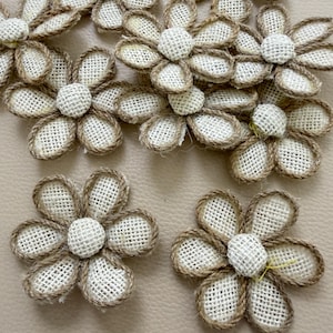 May include: A collection of six-petaled flowers crafted from off-white woven fabric with brown rope-like trim outlining each petal and a circular center piece. The flowers are arranged in a cluster, showcasing the textured material and the consistent design of the individual floral shapes.