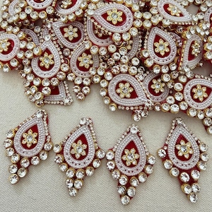 May include: A collection of red, white, and gold decorative embellishments. Each teardrop-shaped piece features a central floral design, surrounded by white beads and clear rhinestones. The embellishments are ideal for craft projects.