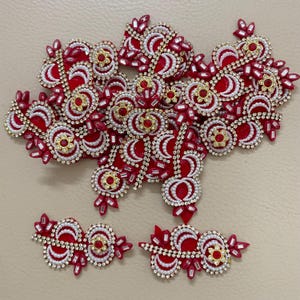 May include: A collection of red, white, and gold decorative embellishments. The embellishments feature intricate designs with beads, sequins, and rhinestones. The pieces are arranged in a scattered pattern, showcasing their ornate details and potential for craft projects.