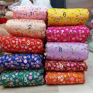 May include: A stack of ten fabric rolls in various colors, including pink, yellow, red, blue, green, and orange. Each roll is adorned with floral embroidery and sequins, creating a textured and decorative appearance. The rolls are stacked vertically, showcasing the variety of colors and patterns.