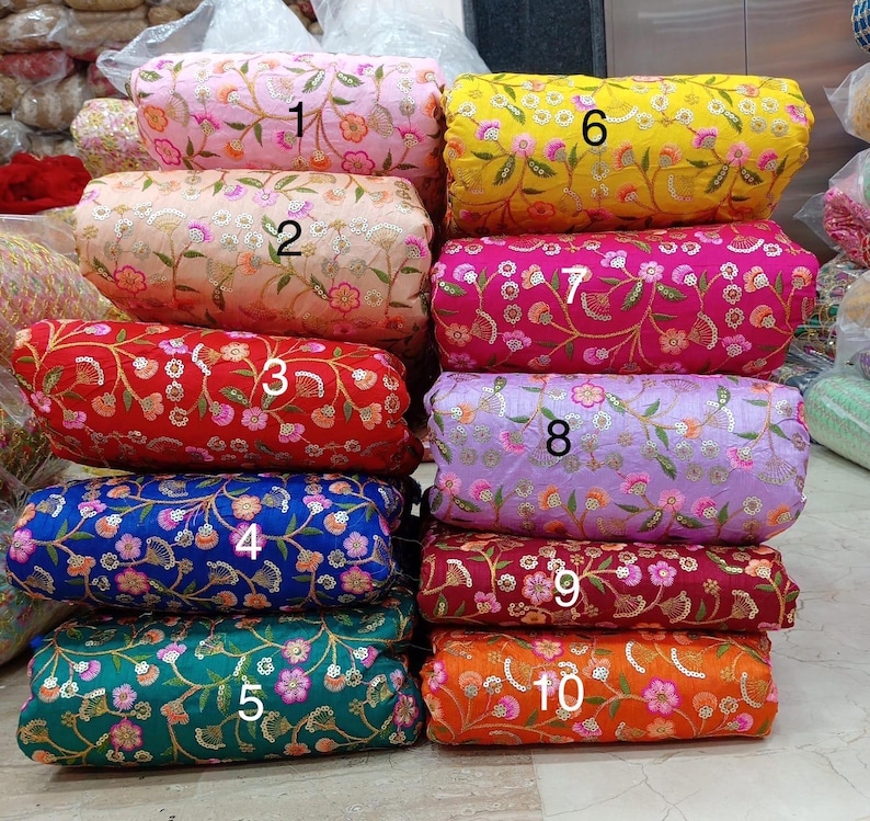 May include: A stack of ten fabric rolls in various colors, including pink, yellow, red, blue, green, and orange. Each roll is adorned with floral embroidery and sequins, creating a textured and decorative appearance. The rolls are stacked vertically, showcasing the variety of colors and patterns.