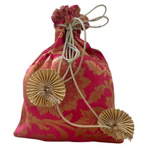 Könnte beinhalten: Ein pinkfarbener Beutel mit Kordelzug, goldenem Blumenmuster und goldenen Zierakzenten. Der Beutel hat zwei goldene fächerförmige Verzierungen und einen Kordelverschluss mit einer goldenen Kordel.