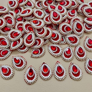 May include: A collection of red teardrop-shaped embellishments. Each piece features a central red gem, surrounded by white pearls, gold trim, and a red base. The embellishments are scattered across a neutral surface.