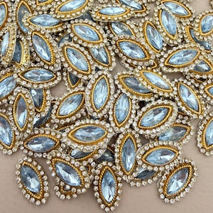 Puede incluir: Una colección de pedrería azul claro en forma de marquesa, cada una engastada en un marco de metal dorado rodeado por un borde de diminutos cristales transparentes. Estos adornos decorativos presentan una piedra central facetada y un detalle de cuerda dorada.