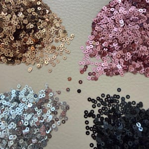May include: An overhead shot displays four piles of sequins in various colors. The colors include gold, rose, silver, and black. The sequins are round and shiny, scattered on a neutral-toned surface. These decorative elements are often used in crafts and fashion.