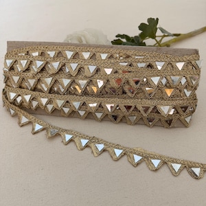 May include: Gold-colored trim with mirrored triangle accents. The trim is wound on a cardboard spool, with a length of the trim unrolled in the foreground. The trim is suitable for embellishing clothing, accessories, or craft projects.