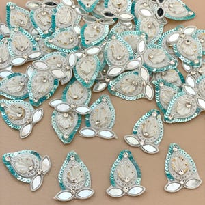 May include: A collection of decorative fabric appliques. Each applique features a central white design with iridescent sequins, small beads, and mirrored accents. The appliques have a teardrop shape with a turquoise border.