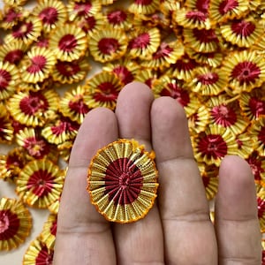 May include: A hand displays a gold and red decorative rosette. The rosette, with a pleated fabric design, is surrounded by similar items. The background is filled with these rosettes, creating a textured, colourful display.