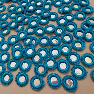Puede incluir: Colección de adornos circulares de color turquesa con un centro de espejo. Cada pieza está rodeada de hilo turquesa. Los adornos están dispersos sobre una superficie neutra, creando un patrón texturizado. Ideal para proyectos de manualidades.
