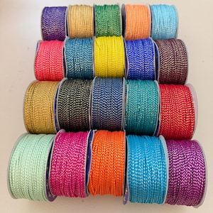 May include: A collection of colorful spools of decorative cord. The spools are arranged in a grid and feature a variety of colors, including purple, gold, green, orange, and blue. The cord has a metallic sheen, adding a touch of sparkle.