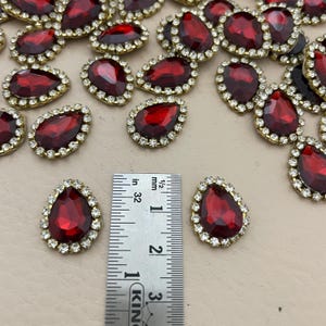 Op de afbeelding: Een verzameling druppelvormige, rode strass-steentjes met een goudkleurige rand en een ring van kleine, heldere strass-steentjes. De strass-steentjes zijn ongeveer 2,5 cm lang. Een liniaal is inbegrepen ter referentie.