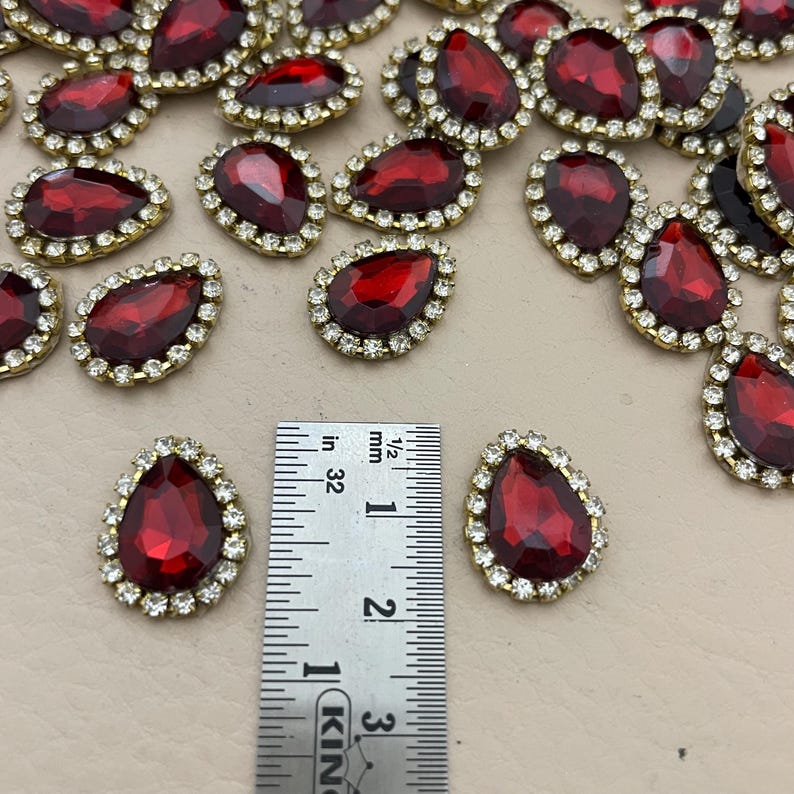 Op de afbeelding: Een verzameling druppelvormige, rode strass-steentjes met een goudkleurige rand en een ring van kleine, heldere strass-steentjes. De strass-steentjes zijn ongeveer 2,5 cm lang. Een liniaal is inbegrepen ter referentie.