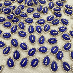 May include: A collection of small, oval-shaped blue cabochons, each featuring a central vertical row of three clear rhinestones and surrounded by a border of tiny, sparkling clear crystals, arranged in a scattered pattern.