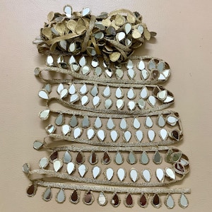 Puede incluir: Ribete decorativo dorado con detalles de espejo reflectantes en forma de lágrima. El ribete está dispuesto en múltiples hebras, con un haz de ribete similar en la parte superior. Los espejos reflejan la luz, creando un efecto brillante. Ideal para adornar prendas o manualidades.