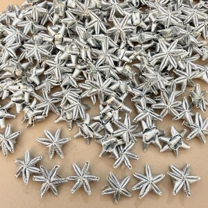 Puede incluir: Un conjunto de adornos plateados en forma de estrella. Estas estrellas decorativas metálicas están dispersas sobre una superficie neutra. Las estrellas tienen un aspecto tejido detallado, perfectas para manualidades y decoraciones festivas.