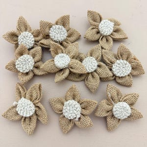 Puede incluir: Un conjunto de ocho flores artesanales hechas de tela de arpillera color canela. Cada flor presenta cinco pétalos redondeados dispuestos en forma de estrella con un centro circular de ganchillo blanco texturizado unido al medio de cada flor.