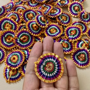 May include: A collection of colorful, circular decorative ornaments. Each ornament features a gold outer ring, with concentric circles of red, green, blue, and white. One ornament is held in a hand, with many more scattered in the background.