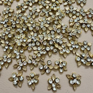 以下が含まれることがあります： 金色の金属製の花の装飾品、透明なクリスタルのアクセント付き。 葉が付いた花の模様で、薄いピンク色の表面に散らばっています。 さまざまなクラフトプロジェクトに適しています。