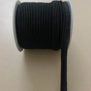 May include: A spool of black piping trim. The trim is tightly wound on a gray plastic spool. A length of the trim extends from the spool, showing its edge. The trim appears to be made of a woven fabric.