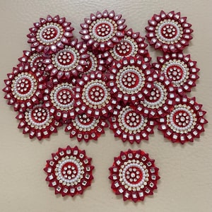May include: A collection of maroon circular embellishments. Each piece features a central design of white pearls and gold accents, surrounded by a ring of small, square, mirrored elements. The outer edge has a petal-like shape.