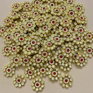May include: A pile of gold-coloured, flower-shaped embellishments. Each piece has a central red gem surrounded by clear, sparkling rhinestones. The petals are outlined with smaller clear rhinestones, creating a dazzling effect. These decorative elements are ideal for crafting and embellishment projects.