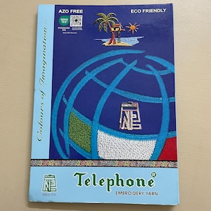 May include: A blue and white Telephone Embroidery Yarn product label. The label features a globe design with embroidered details, a palm tree, and the text "Colours of Imagination." It also includes the text "AZO FREE" and "ECO FRIENDLY."