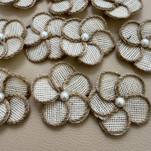 Puede incluir: Una colección de flores de cinco pétalos hechas de tela tejida de color blanco roto con bordes deshilachados. Cada pétalo está delineado con un fino cordel de yute de color natural. Una perla blanca, lisa y redonda, se sitúa en el centro.