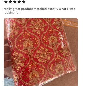 May include: A vibrant red fabric featuring an intricate gold embroidered pattern with floral motifs and small circular sequins. The textile is encased in a clear plastic protective covering, showcasing the detailed metallic thread work throughout.