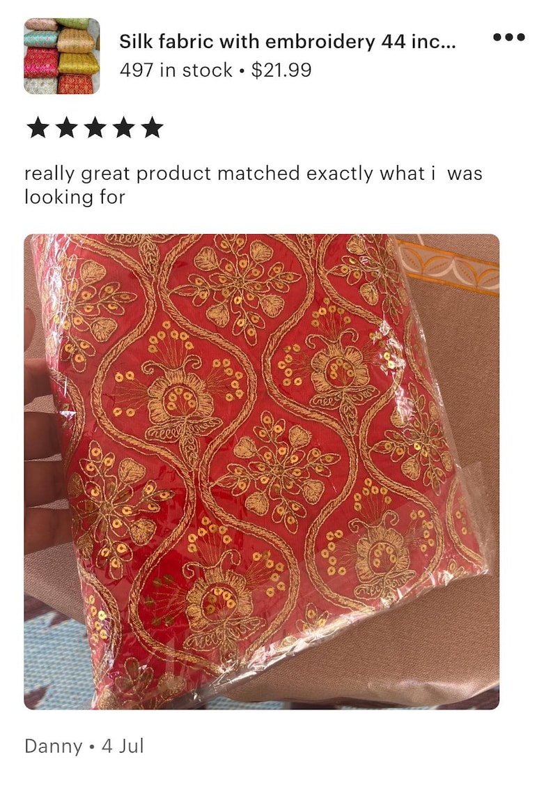 May include: A vibrant red fabric featuring an intricate gold embroidered pattern with floral motifs and small circular sequins. The textile is encased in a clear plastic protective covering, showcasing the detailed metallic thread work throughout.