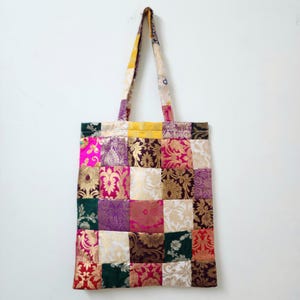 May include: A patchwork tote bag with a variety of colorful fabric squares. The bag features a mix of floral and patterned designs in shades of pink, gold, purple, and green. The handles are made of a coordinating patchwork fabric.