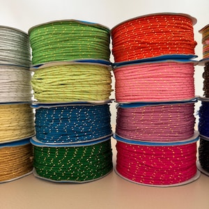 Può includere: Una collezione di rocchetti di cordoncino colorato, ciascuno avvolto ordinatamente su un'anima di cartone. I cordoncini sono in vari colori, tra cui verde, rosso, rosa e blu, con un sottile motivo a macchie dorate. I rocchetti sono impilati.