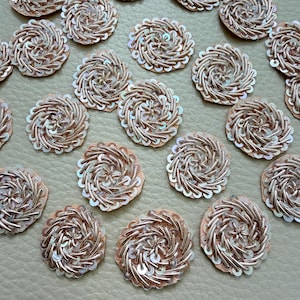 May include: A collection of circular patches featuring intricate rose gold metallic thread embroidery and small clear sequins arranged in a swirling, spiral pattern. Each individual patch has a scalloped edge and a dense, textured surface.