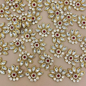 Peut inclure: Gros plan de plusieurs ornements floraux dorés. Chaque fleur a un centre rouge, entouré de cristaux clairs et facettés et de feuilles métalliques dorées. Les ornements sont dispersés sur une surface beige clair.