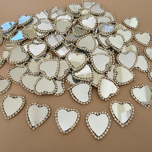 May include: A collection of heart-shaped mirror embellishments. Each mirror has a gold-colored border encrusted with small, clear rhinestones. The mirrors are scattered across a neutral-toned surface, reflecting light and creating a sparkling effect.