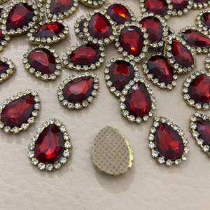 Op de afbeelding: Close-up van talrijke rode, druppelvormige strass-steentjes met een goudkleurige metalen rand en een ring van kleine, heldere strass-steentjes. De strass-steentjes zijn verspreid over een lichtgekleurd oppervlak, waardoor hun glinsterende facetten zichtbaar zijn.