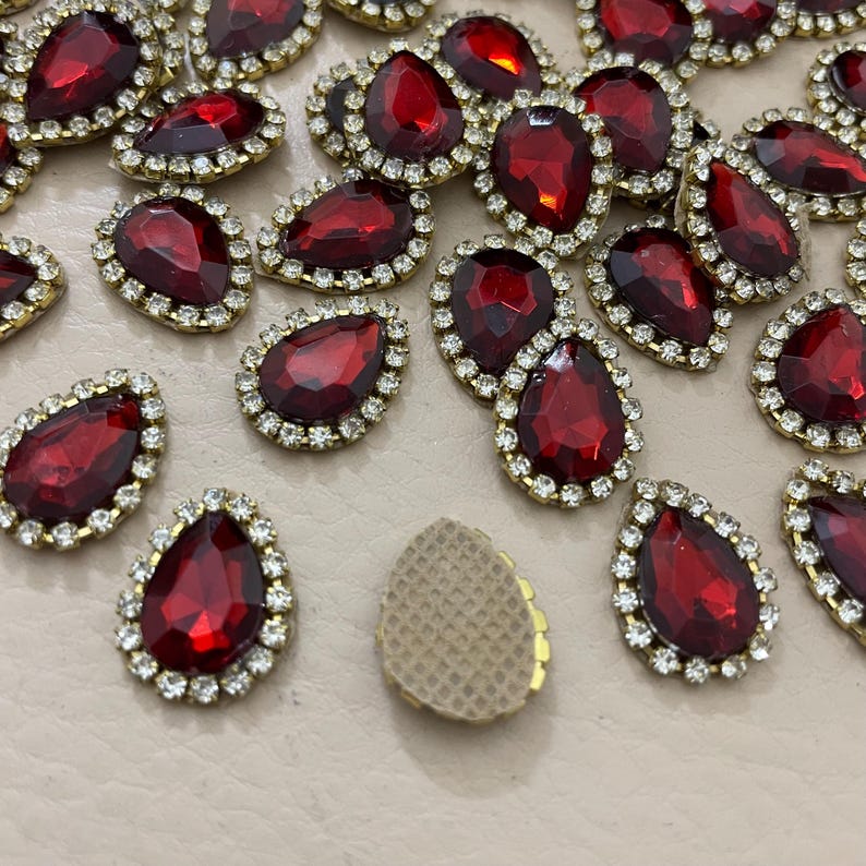 Op de afbeelding: Close-up van talrijke rode, druppelvormige strass-steentjes met een goudkleurige metalen rand en een ring van kleine, heldere strass-steentjes. De strass-steentjes zijn verspreid over een lichtgekleurd oppervlak, waardoor hun glinsterende facetten zichtbaar zijn.