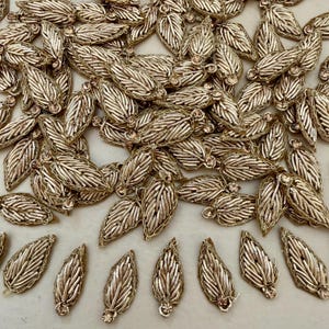 May include: A collection of gold-colored leaf-shaped embellishments. Each leaf is intricately detailed with a woven pattern and a small, clear gemstone at the base. The leaves are scattered across a light-colored surface, creating a decorative display.