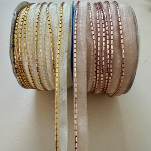 Pode incluir: Duas bobinas de acabamento decorativo, uma com detalhes de contas douradas e outra com detalhes de contas em ouro rosa. O acabamento está em uma fita transparente de cor branco sujo. Ideal para projetos de costura e artesanato.
