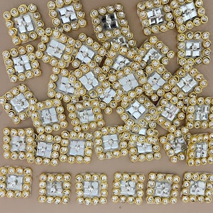 May include: A collection of square embellishments. Each square features a central, clear, faceted gem surrounded by smaller, clear gems and a gold-coloured border. The embellishments are scattered across a neutral surface.