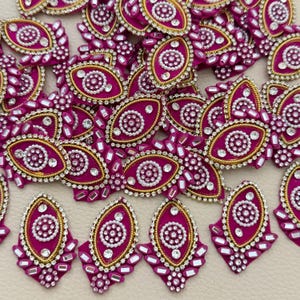 Puede incluir: Una colección de parches en forma de ojo de tela magenta vibrante, cada uno con un patrón circular central de cuentas blancas, bordes de hilo metálico dorado y cristales transparentes en un diseño ovalado y puntiagudo.