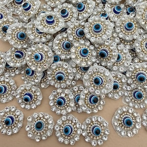 May include: A collection of circular patches featuring a central blue, white, and black evil eye motif surrounded by a ring of small white beads and an outer border of clear, faceted rhinestones set against a white fabric backing.