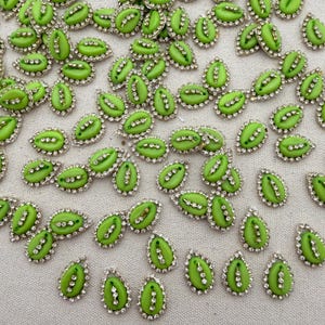 Pode incluir: Uma coleção de pequenos cabochões verde-limão em forma de lágrima, cada um contornado por uma fina armação de metal prateado incrustada com minúsculos strass transparentes. O centro de cada peça apresenta uma fila vertical de três pequenos strass.