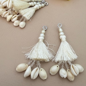 May include: Ivory-colored tassel earrings with a cluster of small seashells. The earrings feature a beaded top, a tassel, and a collection of cowrie shells. The design is suitable for jewelry making or as decorative accents.