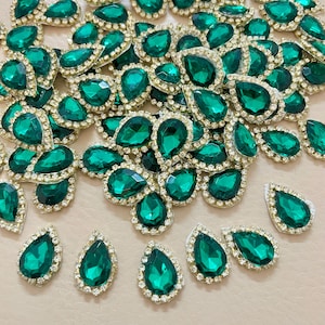 May include: A collection of emerald green teardrop-shaped rhinestones with a gold-colored, rhinestone-studded border. The gemstones are scattered across a light-colored surface, showcasing their faceted surfaces and reflective qualities. These embellishments are ideal for crafting and jewelry making.